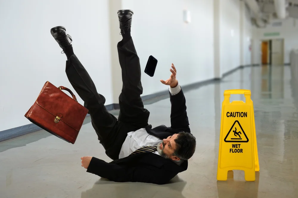 Man slipping and falling on a wet hard floor beside a caution sign